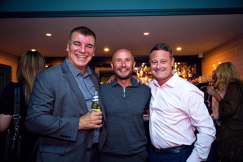 Three men standing in a row holding drinks and smiling